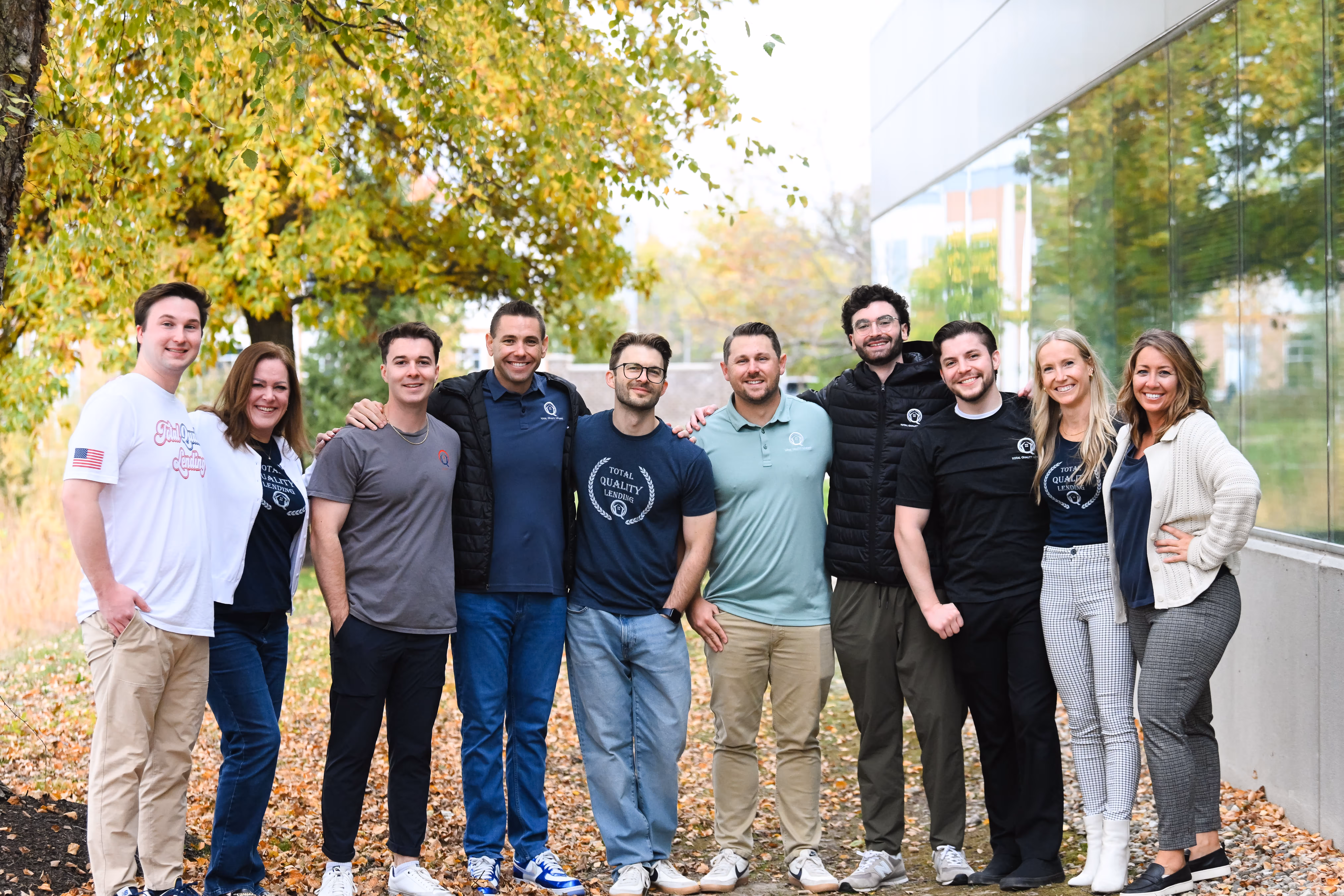 Group of ten people standing closely outdoors in front of a tree and a building with glass windows, smiling and casually dressed in autumn. - Total Quality Lending Images