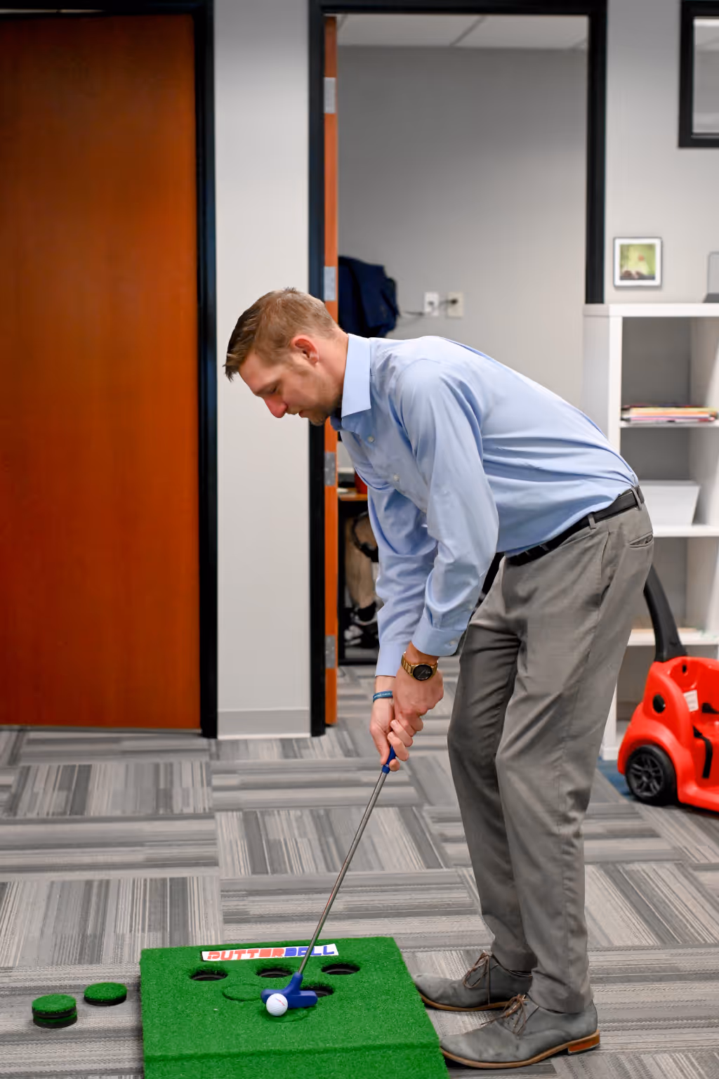 Man in business attire practicing putting on a green indoor putting mat labeled 'PUTTERBALL' in an office setting. - Total Quality Lending Images