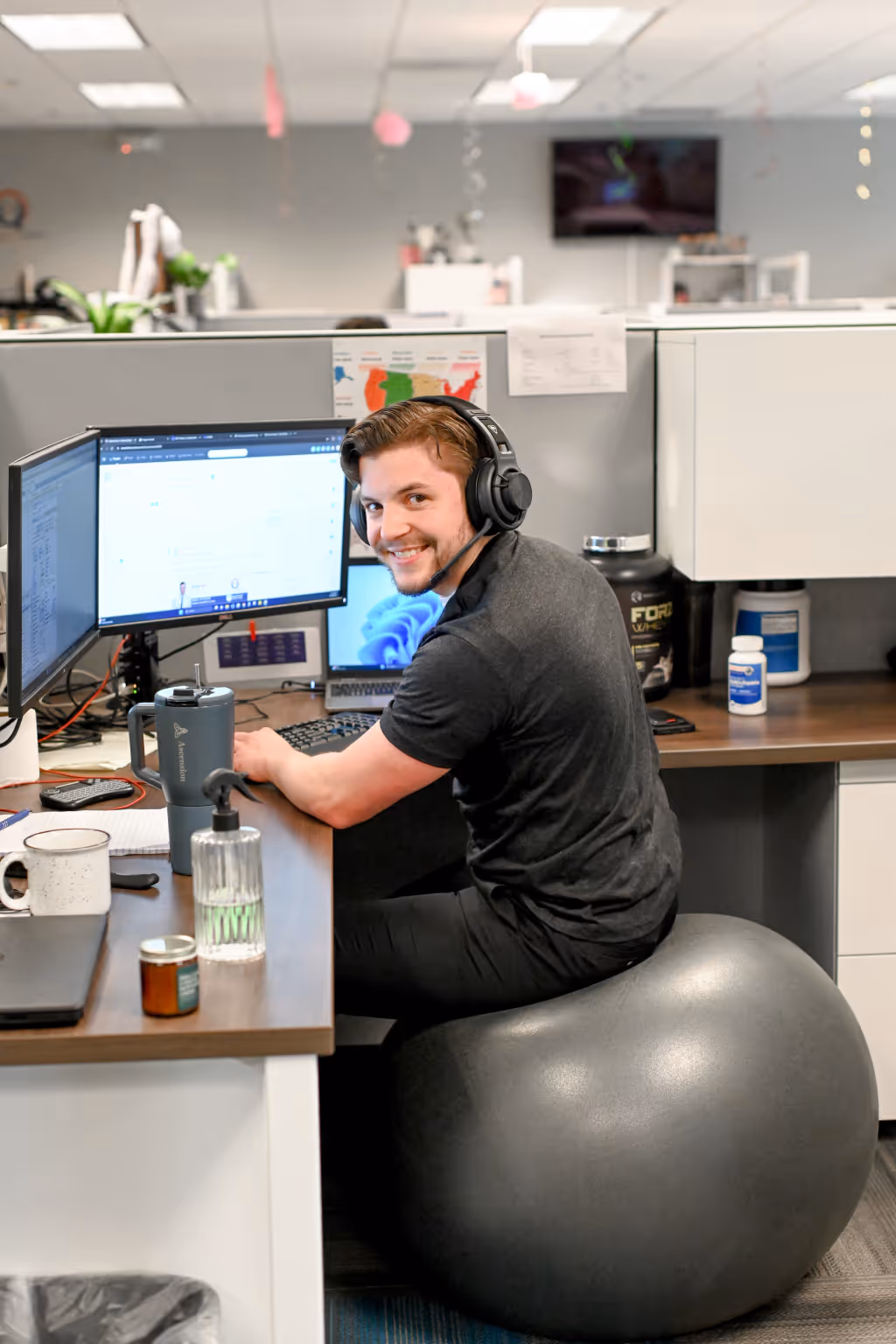 Young man wearing a headset sitting on a gray exercise ball at a cubicle desk with dual monitors, smiling at the camera. - Total Quality Lending Images