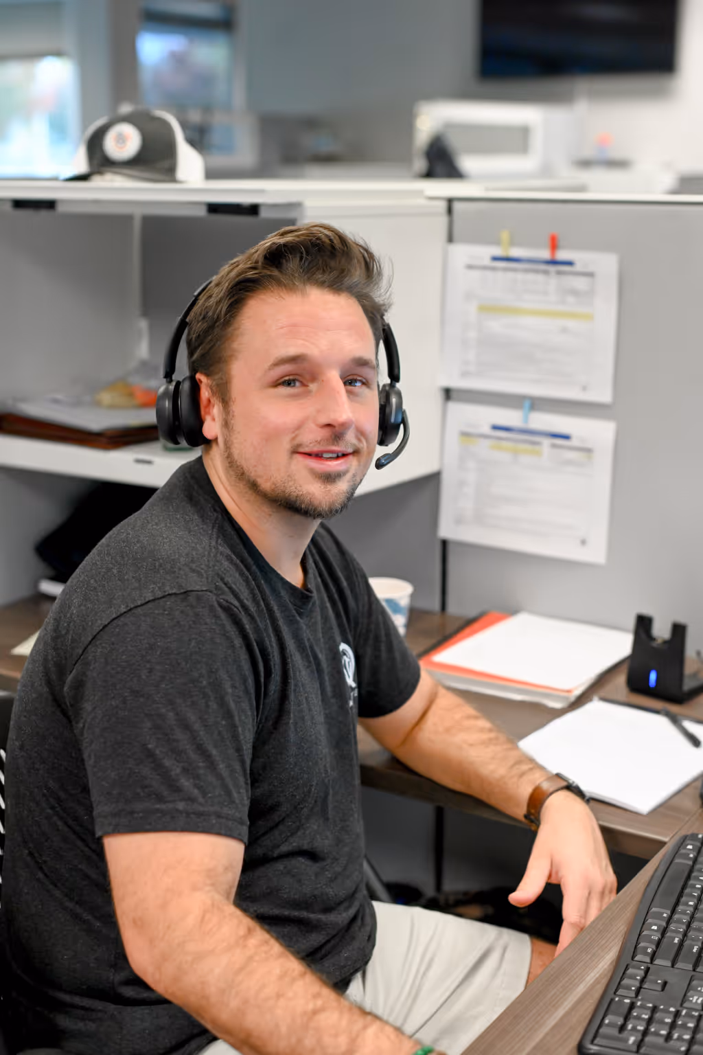 Young man wearing a black headset and dark t-shirt sits at a desk with a keyboard, documents, and a coffee cup in an office cubicle. - Total Quality Lending Images