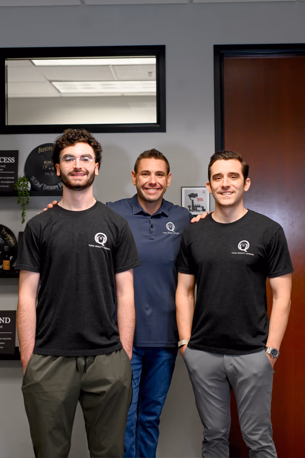 Three men smiling and standing indoors, two wearing black Total Quality Lending t-shirts and one wearing a navy polo shirt with the same logo. - Total Quality Lending Images