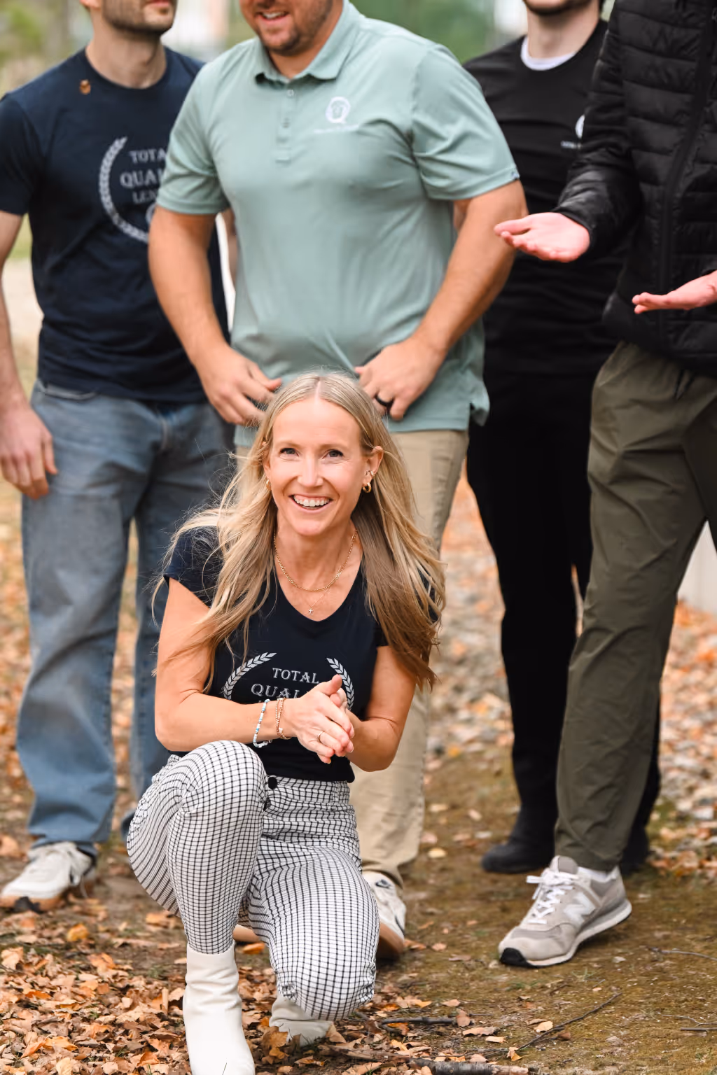 Smiling woman kneeling on a leaf-covered path with men standing behind her in casual outdoor clothing. - Total Quality Lending Images