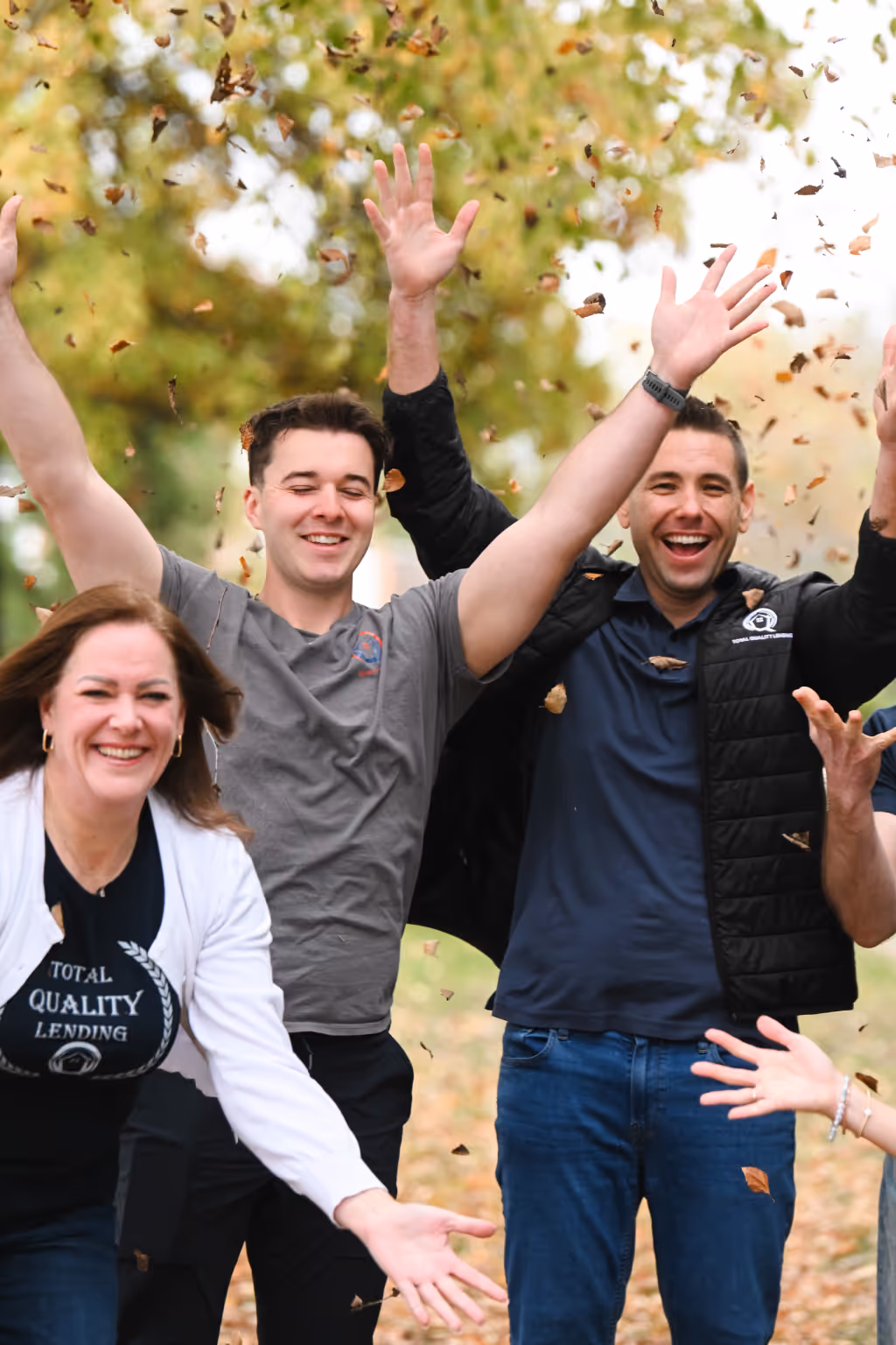 Three adults smiling and joyfully tossing autumn leaves in the air outdoors. - Total Quality Lending Images