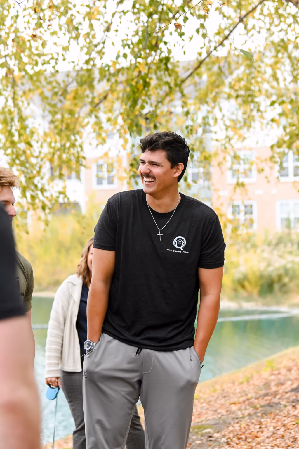 Young man smiling and standing outdoors with hands in pockets, a pond and autumn trees in the background. - Total Quality Lending Images