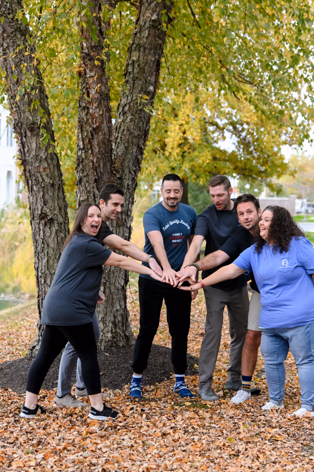 Six people standing outdoors in autumn with hands stacked together in a team gesture under large trees with fallen leaves on the ground. - Total Quality Lending Images