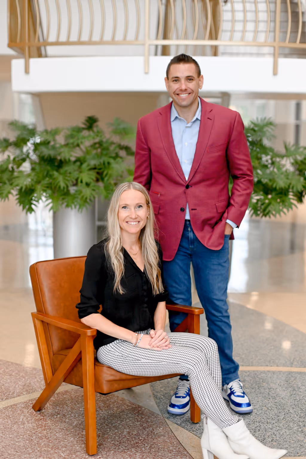 Smiling man in a red blazer and woman in a black blouse and checkered pants posing indoors with green plants in the background. - Total Quality Lending Images