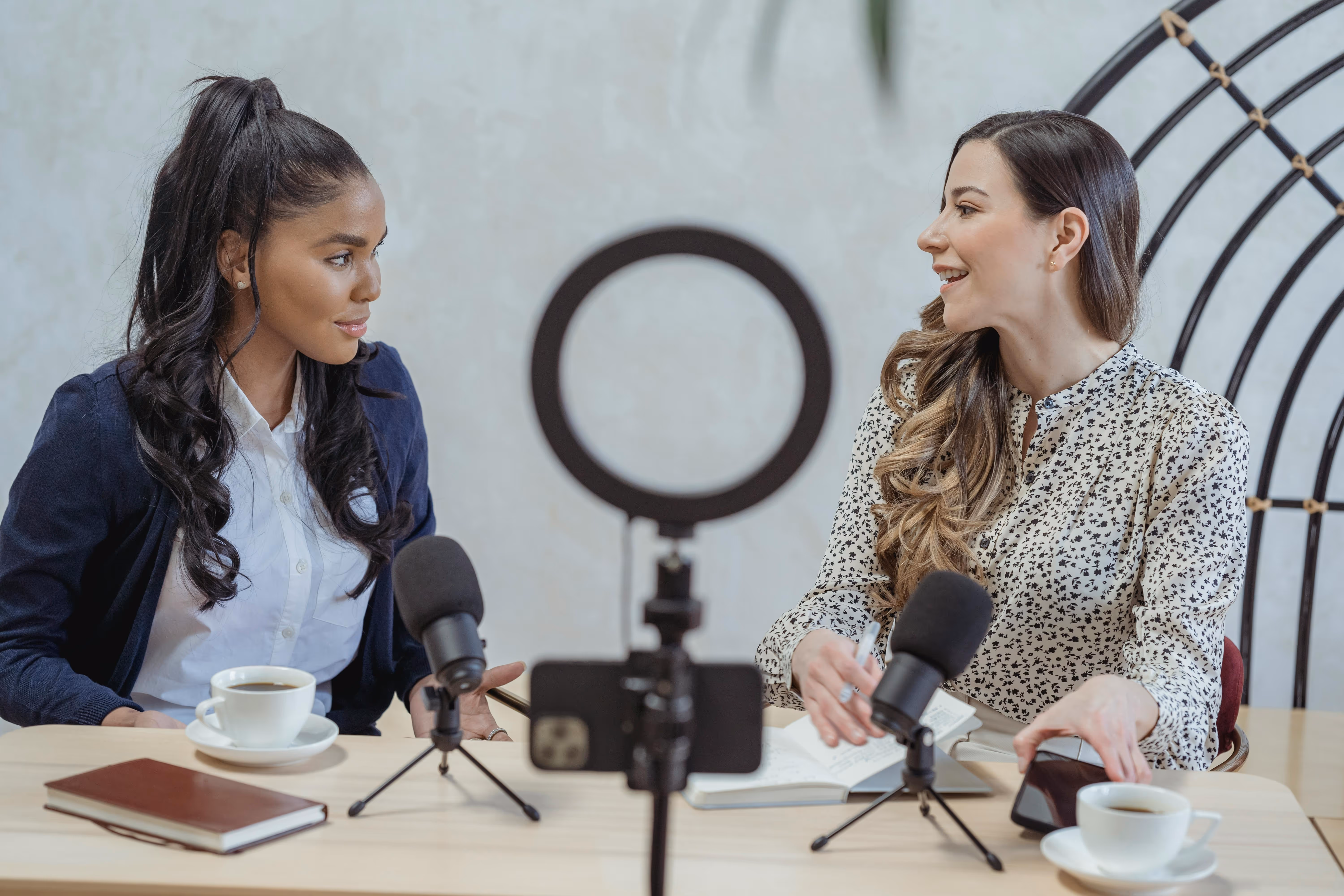 Two women sitting at a table, recording a podcast with microphones, a phone on a tripod, and cups of coffee. - Total Quality Lending Images