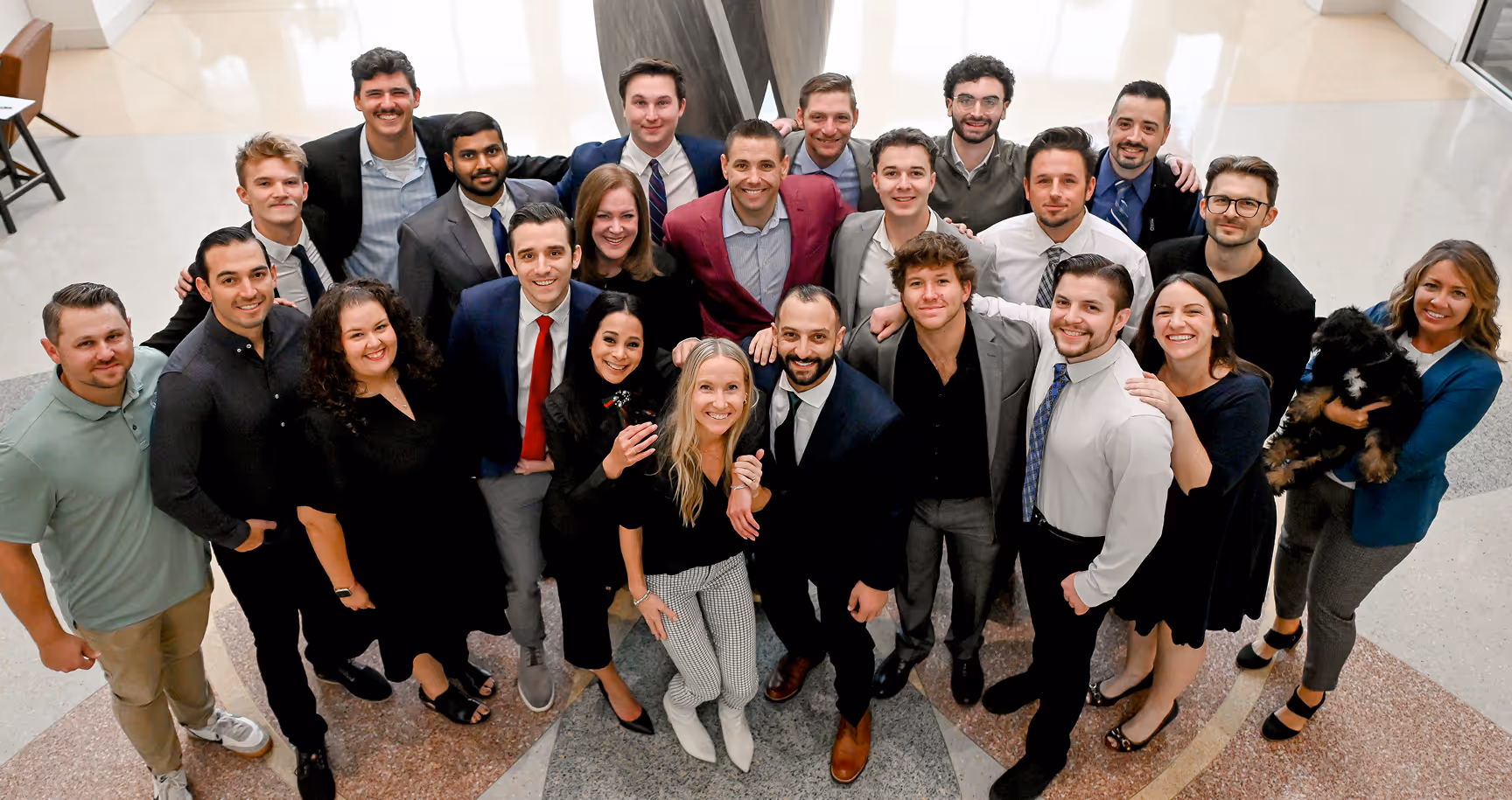 Group of 24 diverse professionals smiling and posing in a modern lobby, one person holding a small black dog. - Total Quality Lending Images