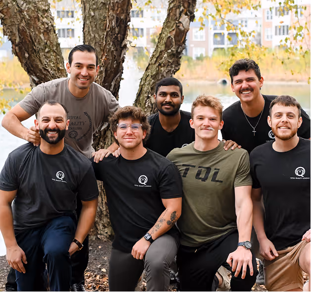 Group of seven smiling young men outdoors in front of trees and buildings, wearing casual T-shirts, some with logos. - Total Quality Lending Images