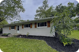 Single-story white brick house with brown shutters surrounded by green grass and trees under a cloudy sky. - Total Quality Lending Images