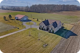 A large house with a dark roof and landscaped yard next to a detached garage on a spacious rural property with fields and trees. - Total Quality Lending Images