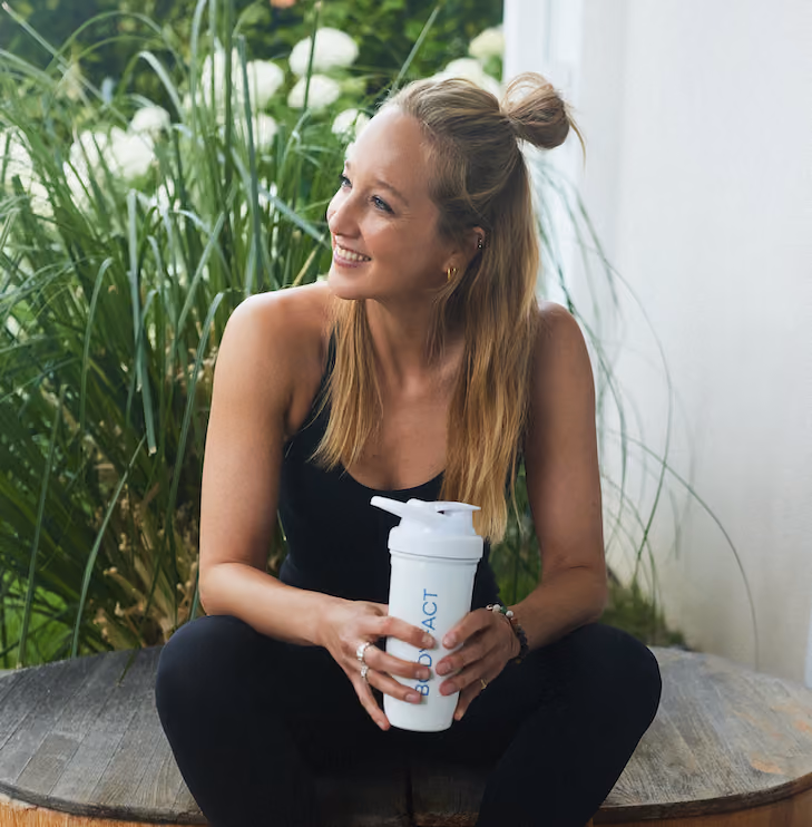 Lächelnde Frau sitzt im Freien und hält eine BODYFACT Shaker-Flasche.
