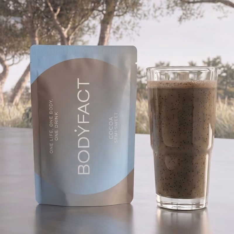 BODYFACT Cocoa Semi-Sweet pouch next to a glass of shake, with an outdoor background.