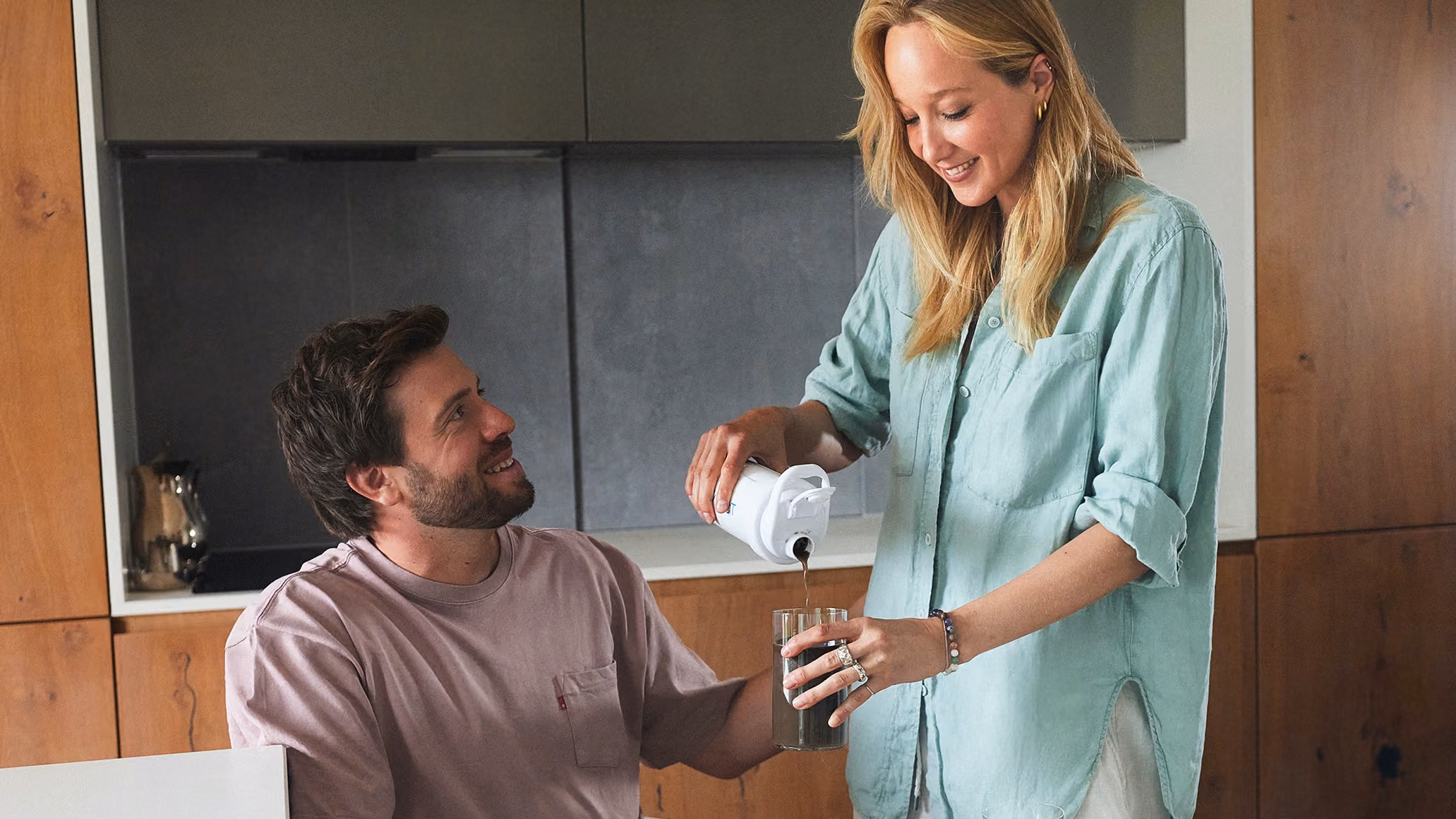 Woman pouring a drink from a BODYFACT shaker bottle into a glass, with a smiling man seated beside her.