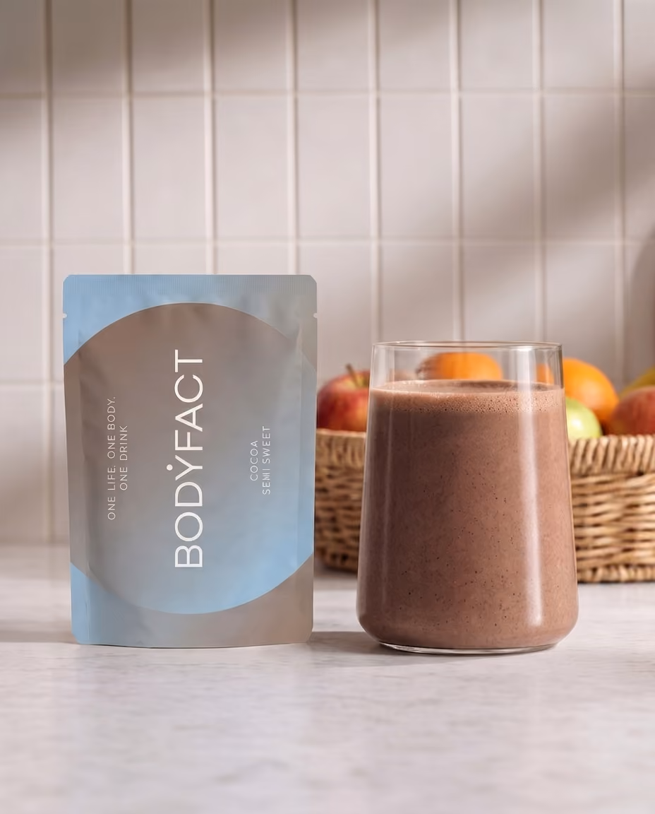 BODYFACT Cocoa Semi Sweet tasak egy pohár kakaós ital mellett, háttérben gyümölcsöskosárral.