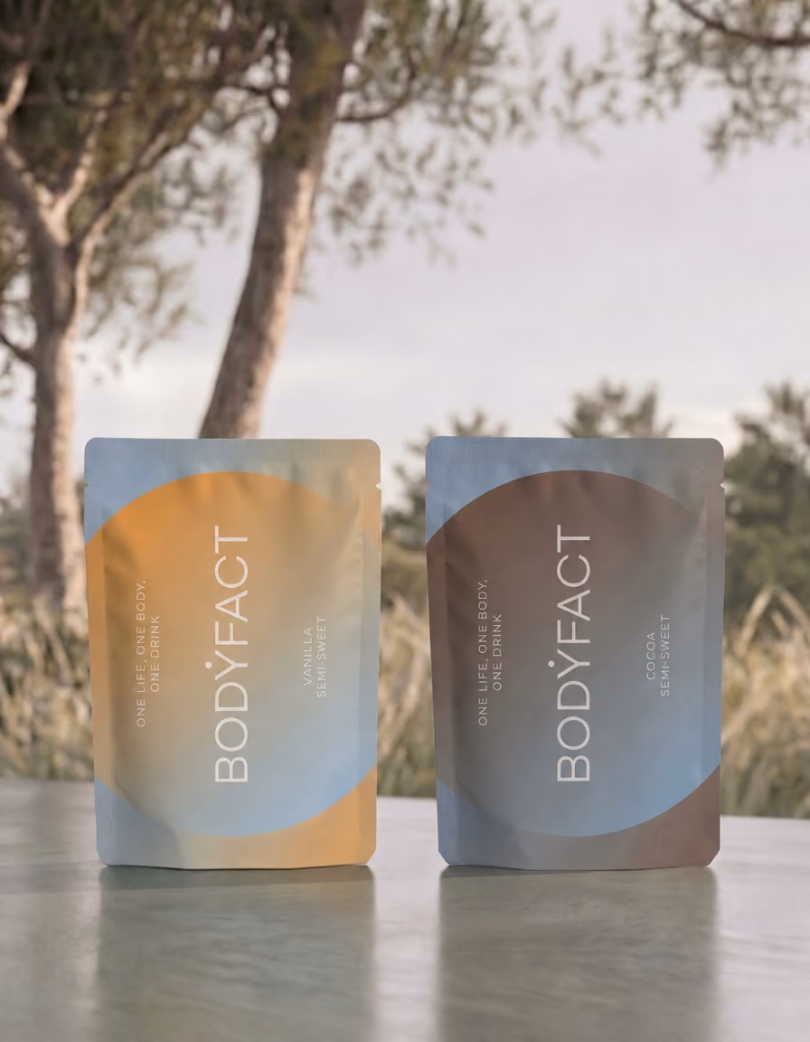 Két BODYFACT tasak, Vanilla és Cocoa Semi Sweet, kültéren, fák előtt.