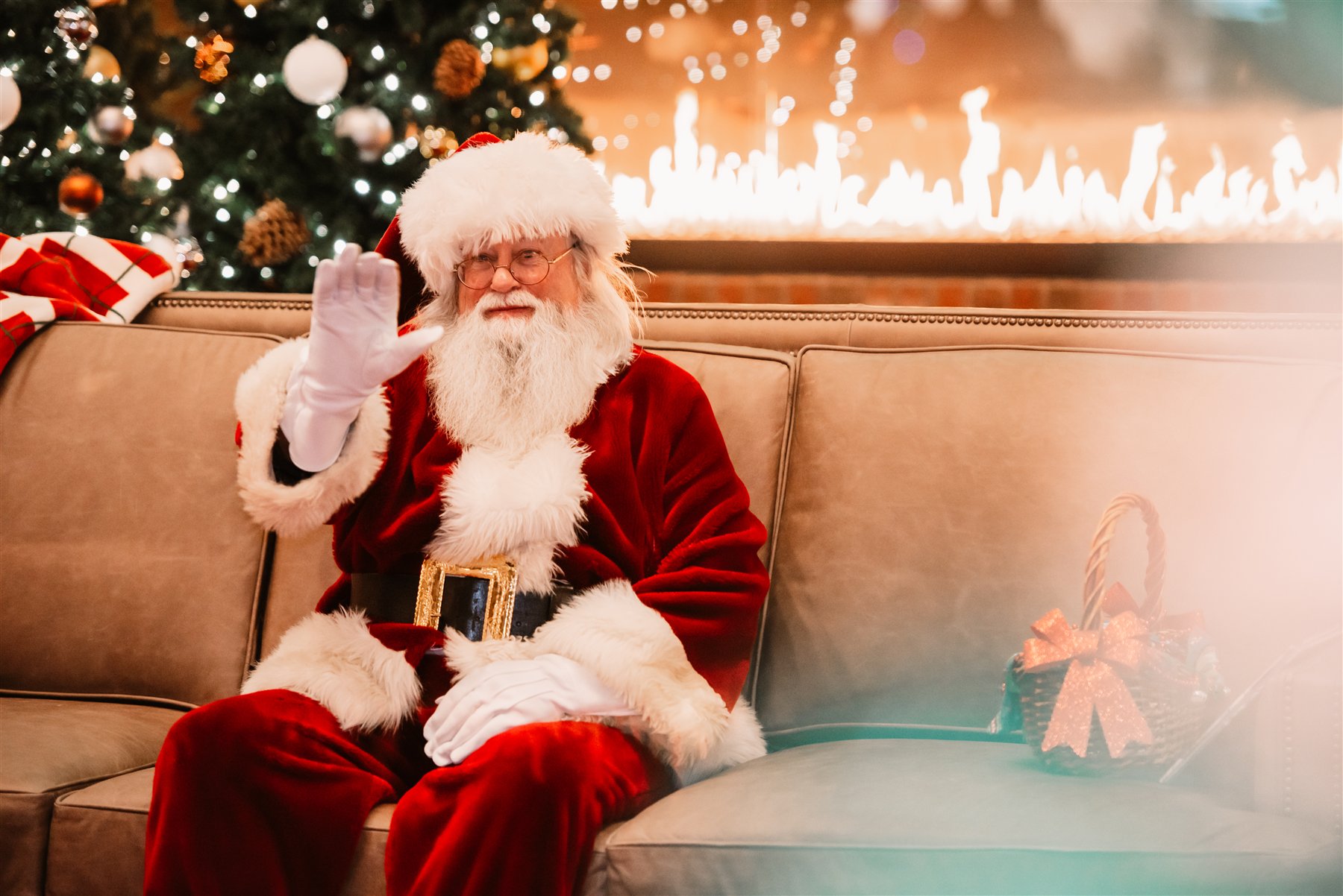 Santa Marty waving from a sofa beside a warmly lit Christmas fireplace