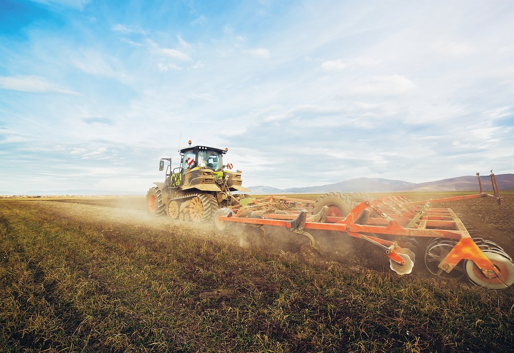 A tractor pulls a disc harrow across a dry field under a bright sky, raising dust.