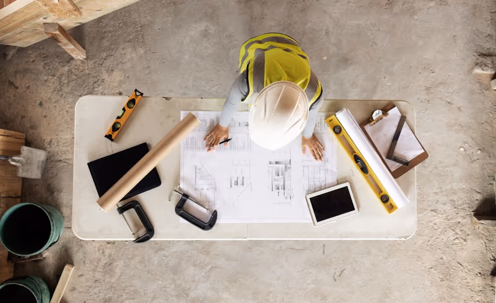 Construction worker in a white hard hat and safety vest reviewing blueprints on a worktable with tools and a tablet.