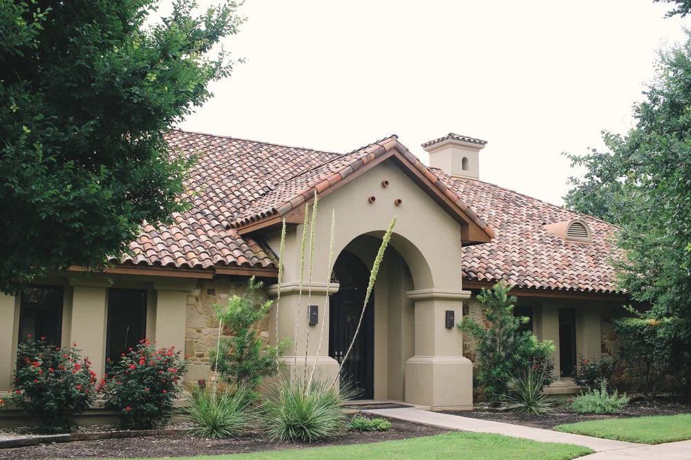Single-story house with beige stucco walls, red tile roof, arched entryway, and landscaped front yard with shrubs and trees.