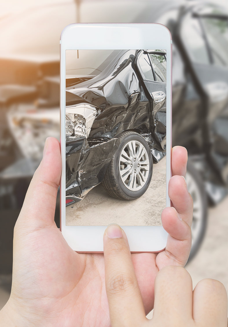 Person holding a smartphone showing a photo of the damaged rear side of a black car with a large dent above the rear wheel.