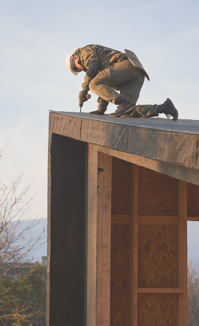 Construction worker wearing a helmet and protective gear kneeling on a roof while using a drill.