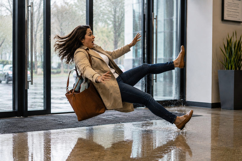 Woman in coat slipping on a wet floor inside a building near glass doors with arms flailing.