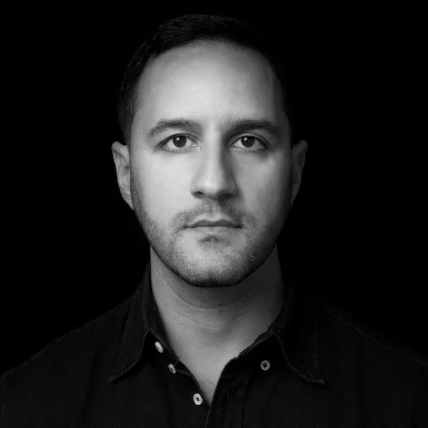 Black and white portrait of a man with short hair and stubble wearing a dark button-up shirt against a black background.