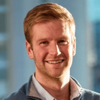 Smiling man with light brown hair wearing a blue jacket and white shirt against a blurred indoor background.