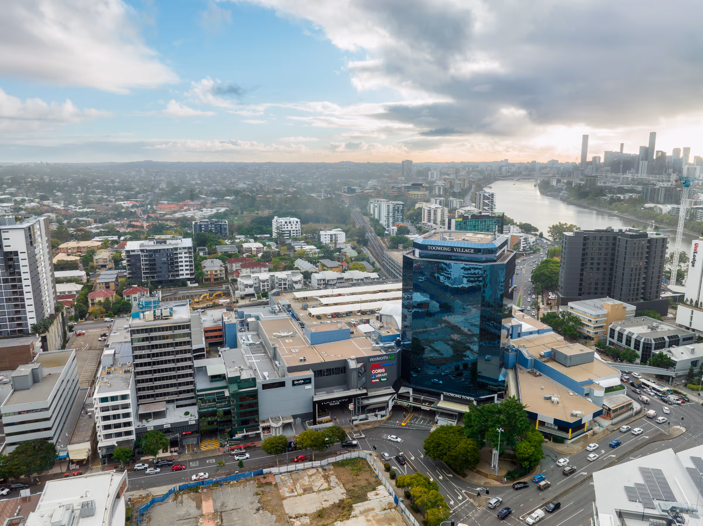 Aerial view of Toowong Tower and Toowong Village shopping center beside a river in a city with surrounding buildings and cloudy sky.