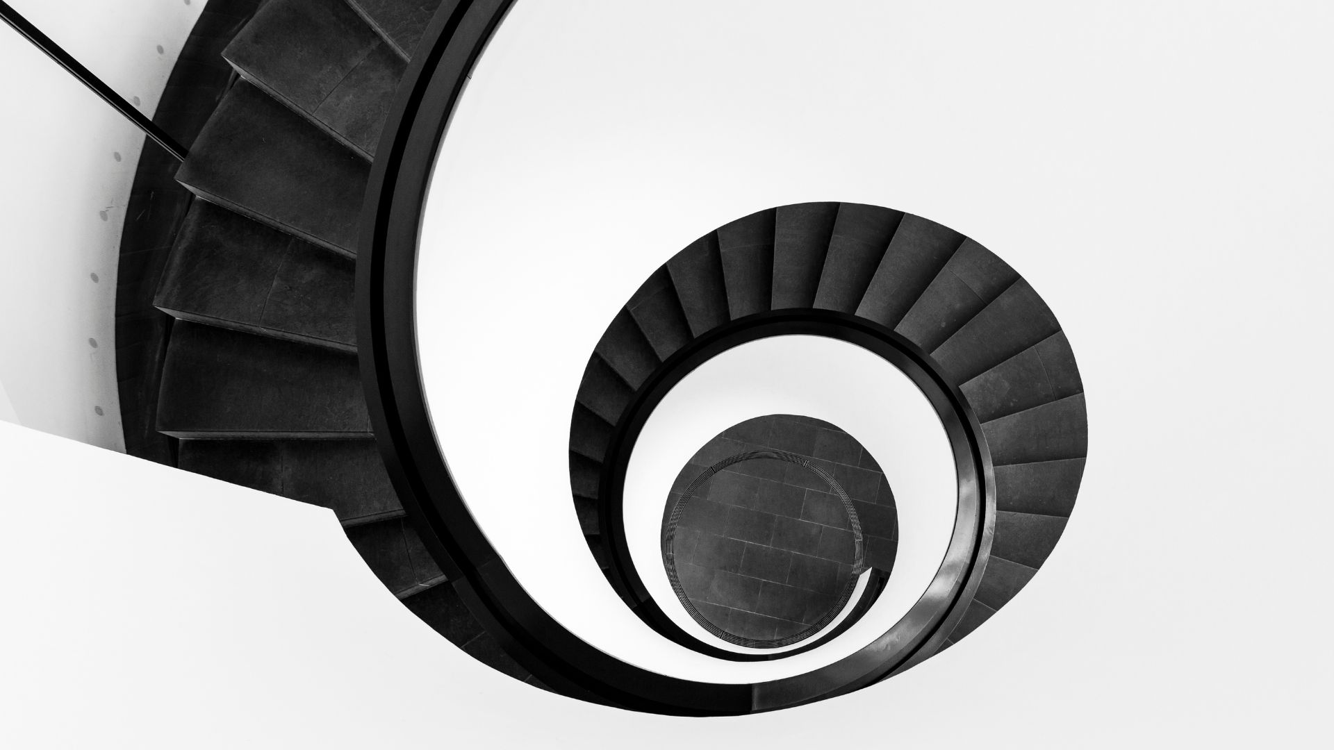 Scala a chiocciola vista dall'alto con gradini scuri e ringhiera nera su sfondo bianco.