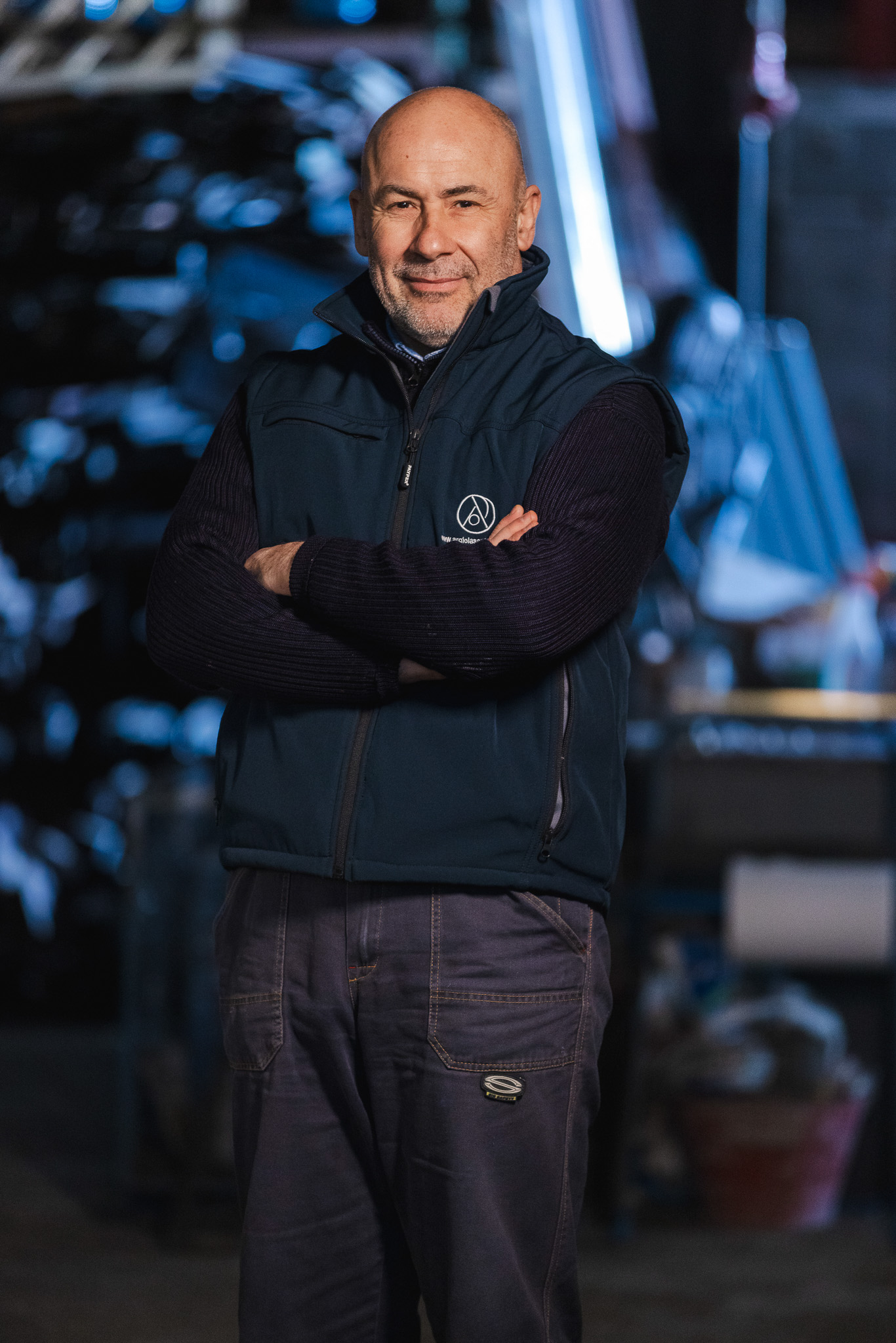 Uomo sorridente con gilet blu e maglione nero in un ambiente industriale.
