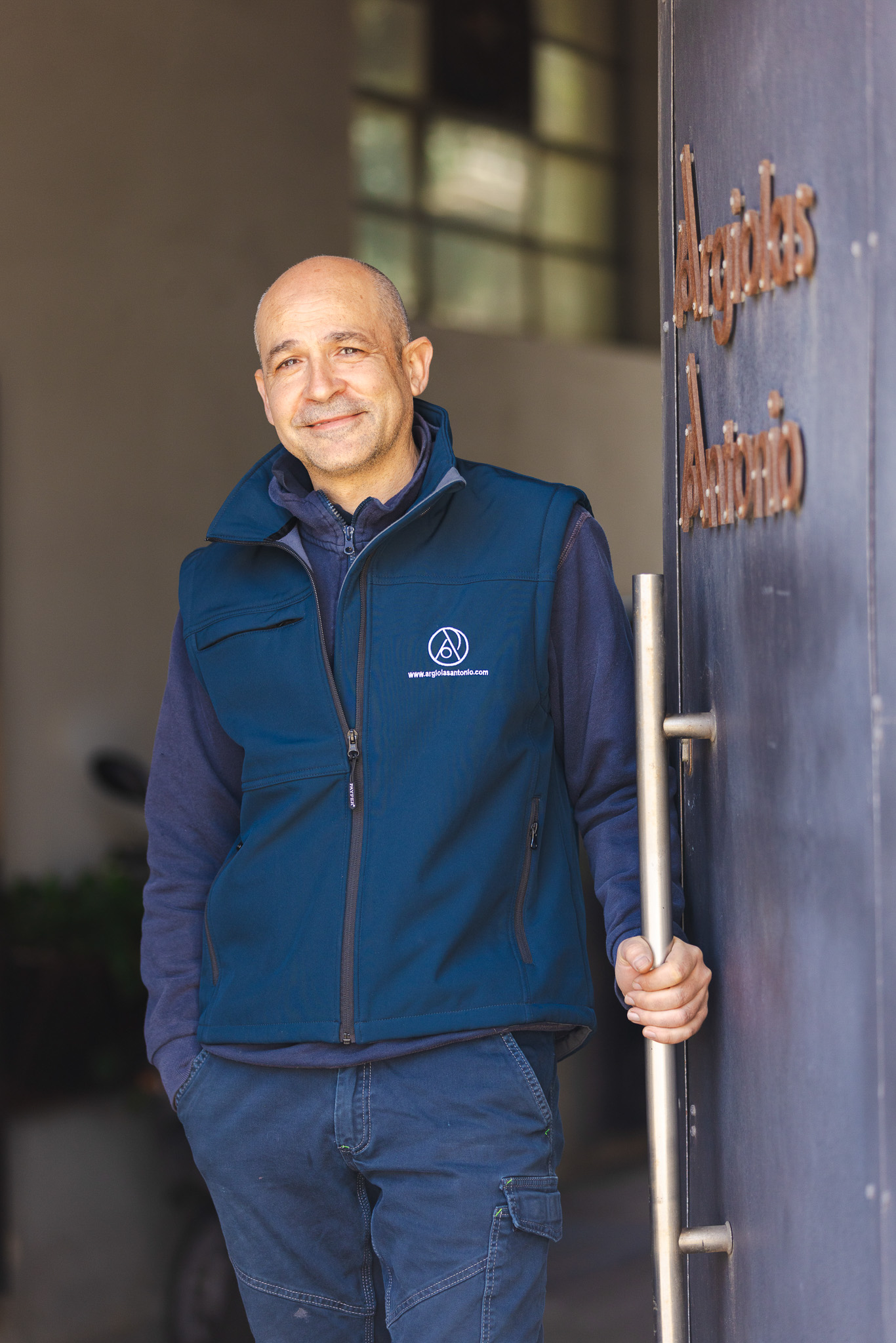 Uomo calvo sorridente con giubbotto blu e pantaloni cargo che tiene la maniglia di una porta con scritto Argolas Antonio.