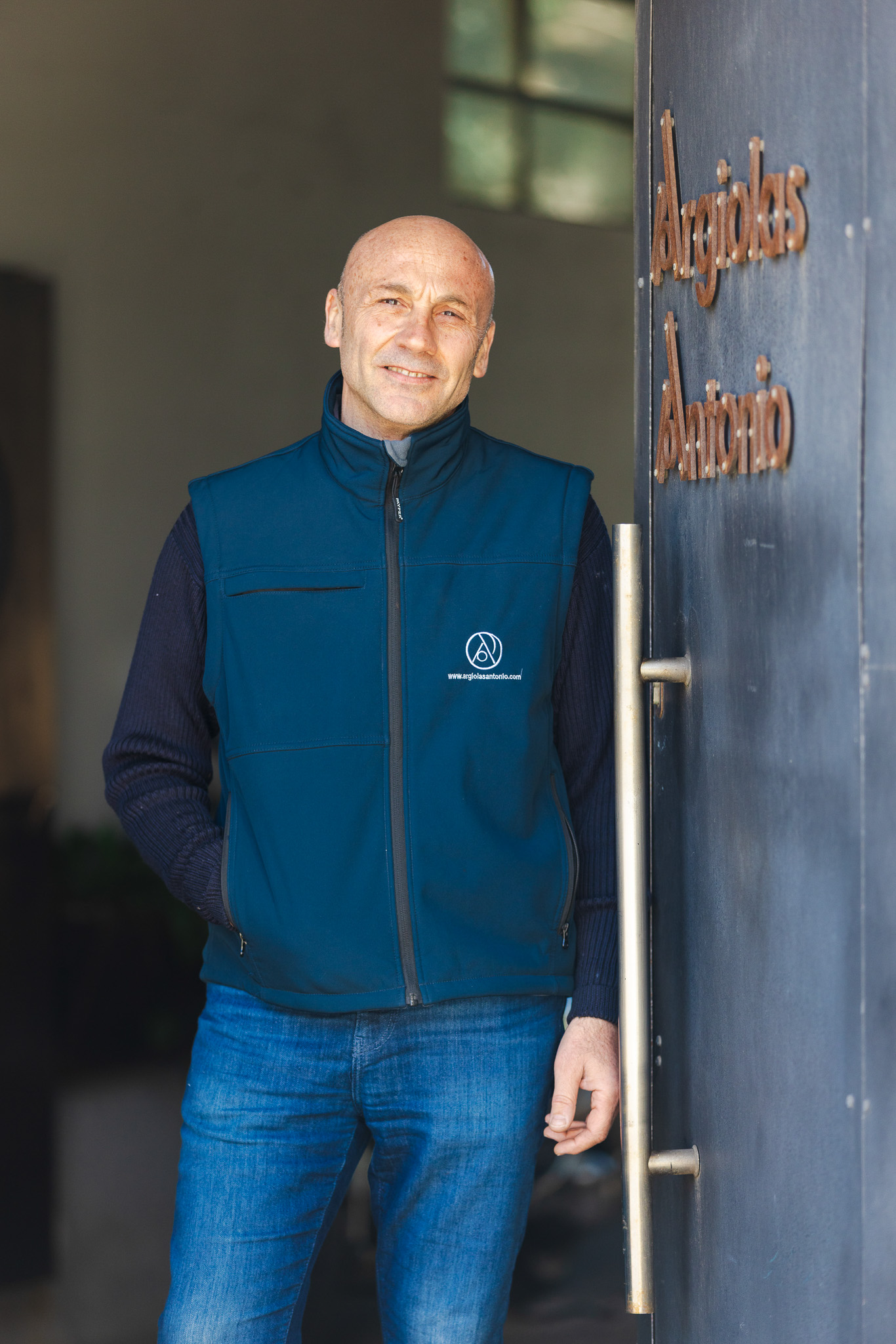 Uomo calvo con gilet blu e jeans vicino a una porta con la scritta Argolas Antonio.