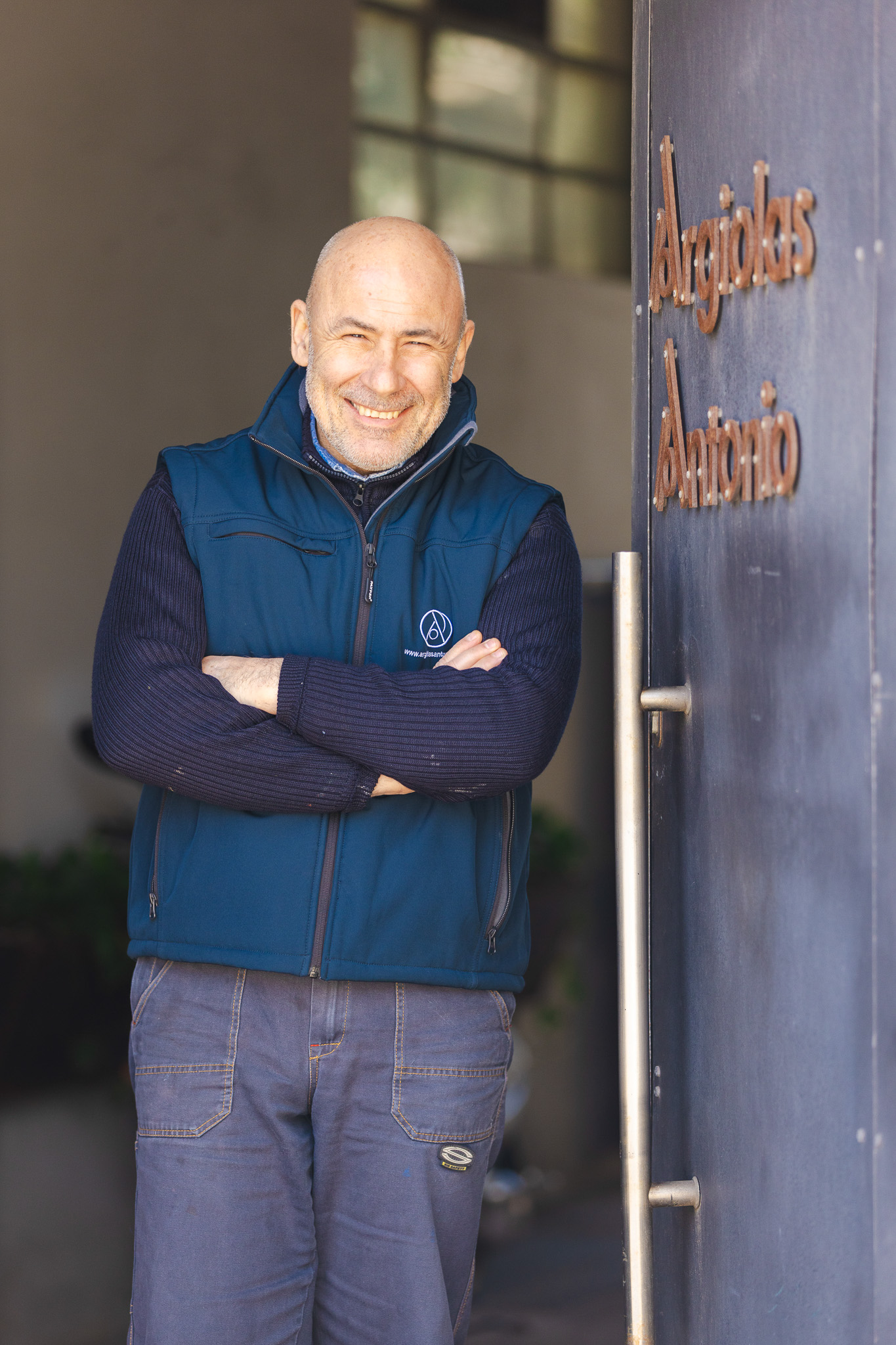 Uomo calvo sorridente con gilet blu e maglione con le braccia incrociate, in piedi accanto a una porta con insegna 'Argiolas Antonio'.