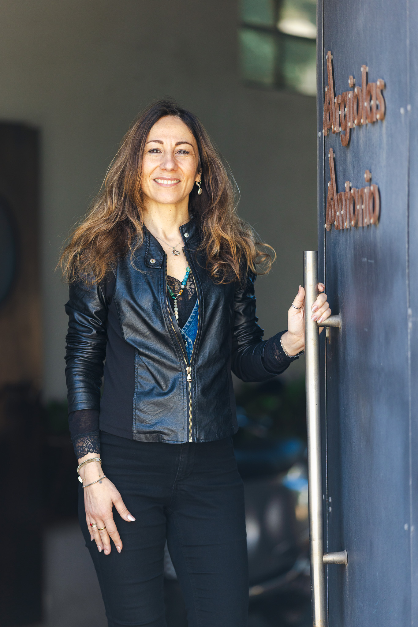 Donna sorridente con giacca di pelle nera che apre una porta scura con maniglia metallica.