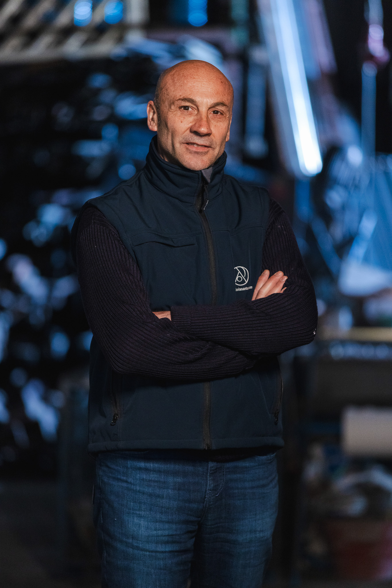 Uomo calvo con gilet blu scuro e maglione nero, braccia incrociate, in un ambiente interno con luci soffuse sullo sfondo.