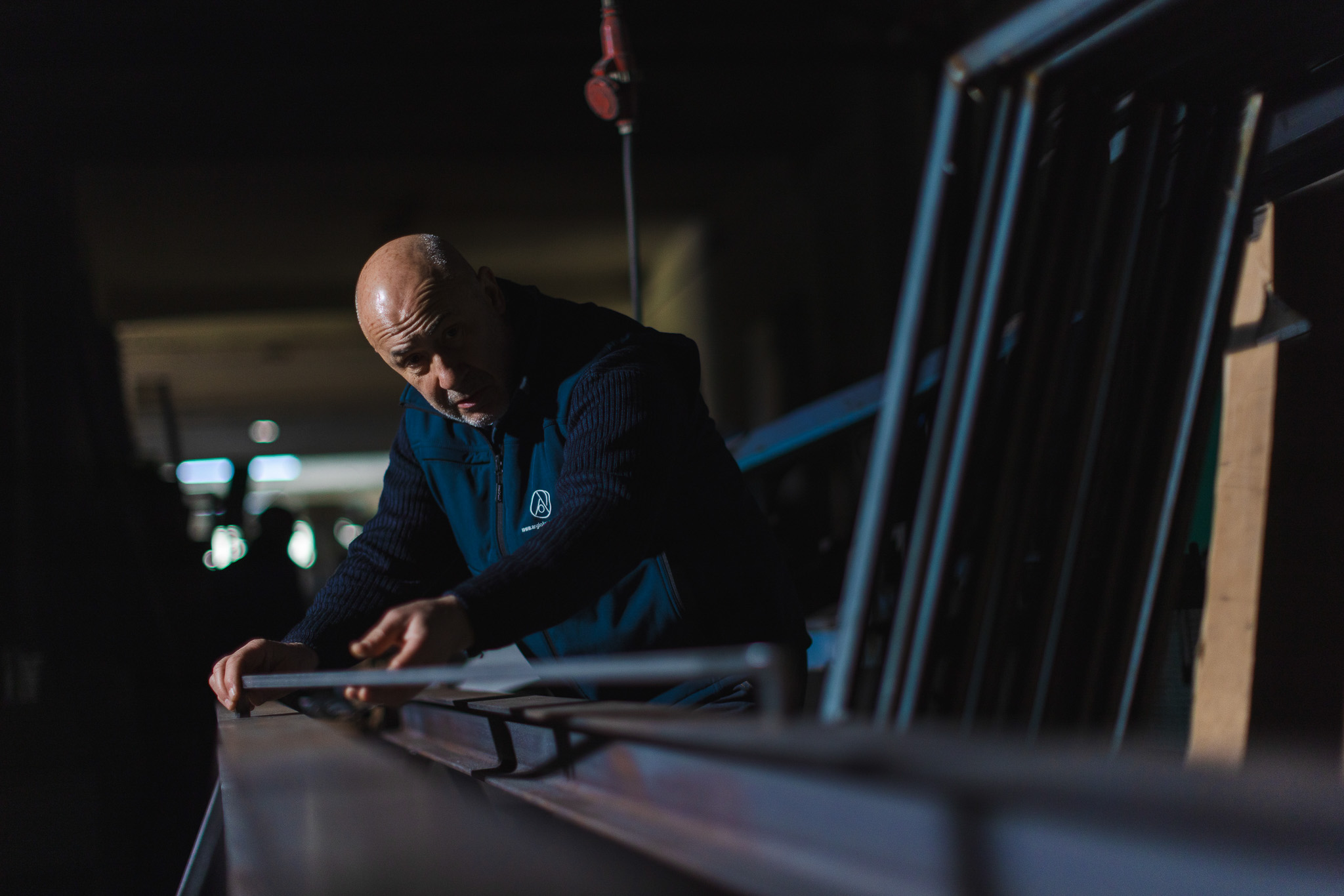 Uomo calvo con giacca scura che lavora con utensile su una superficie in legno in un laboratorio poco illuminato.