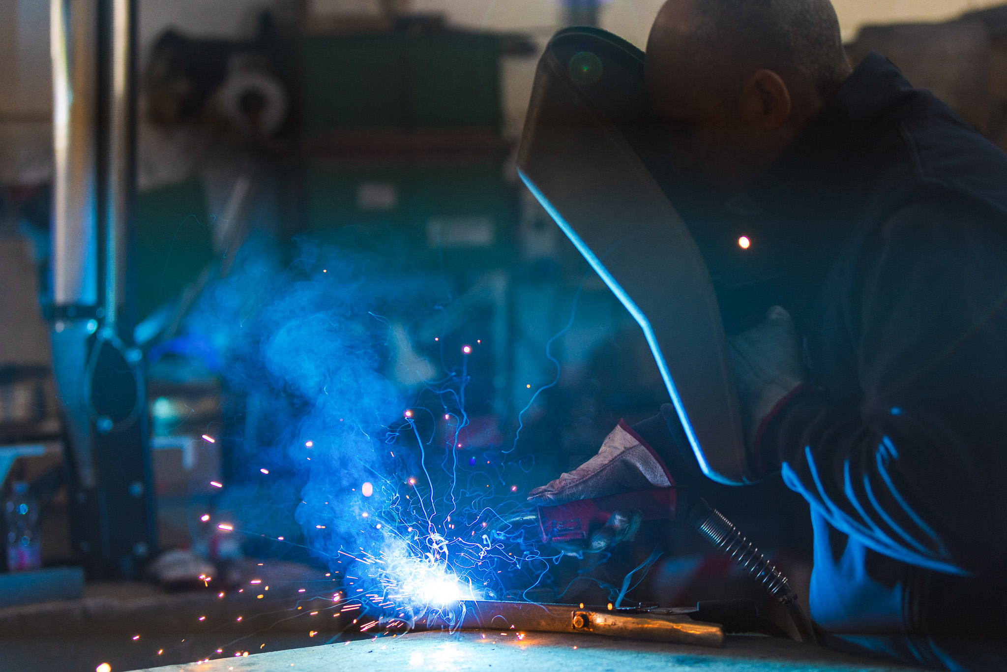 Operaio con maschera da saldatura che esegue una saldatura con scintille e fumo blu in officina.
