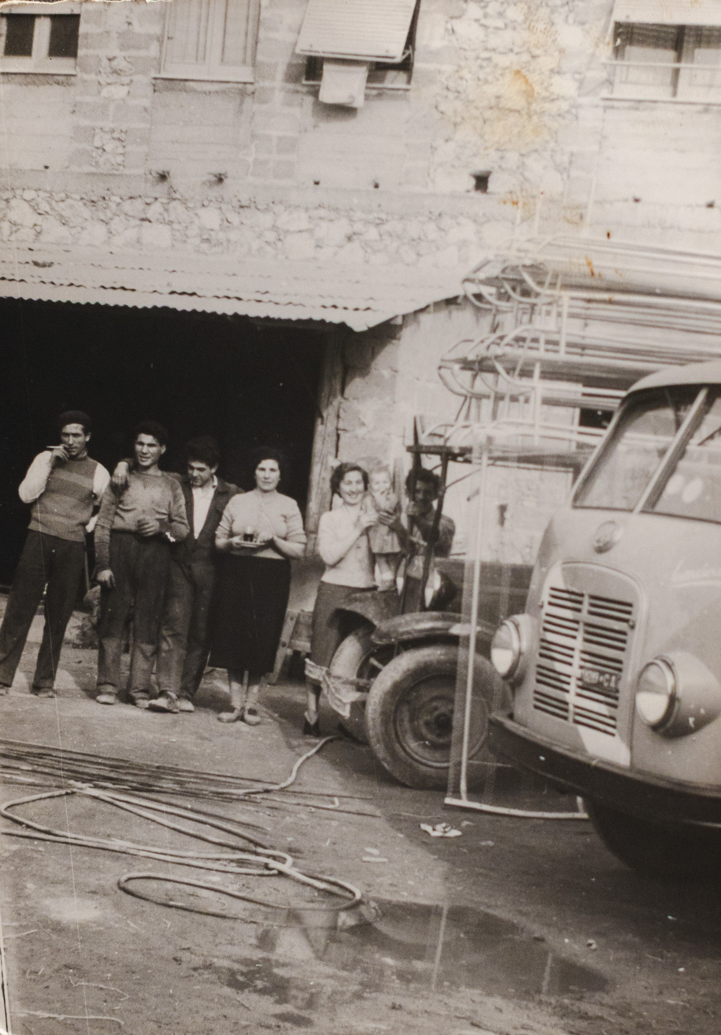 Gruppo di sette persone in abiti d'epoca davanti a un edificio di pietra con un camion e attrezzi da lavoro.