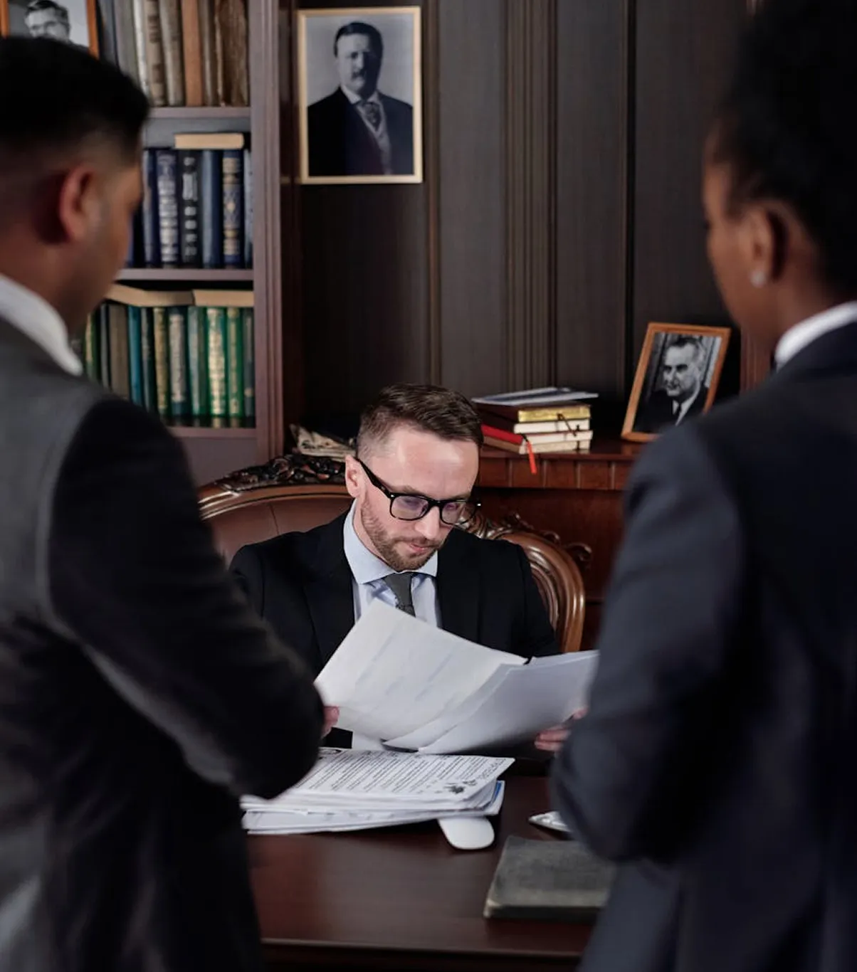 Man in glasses reading through papers at a desk while two colleagues stand nearby
