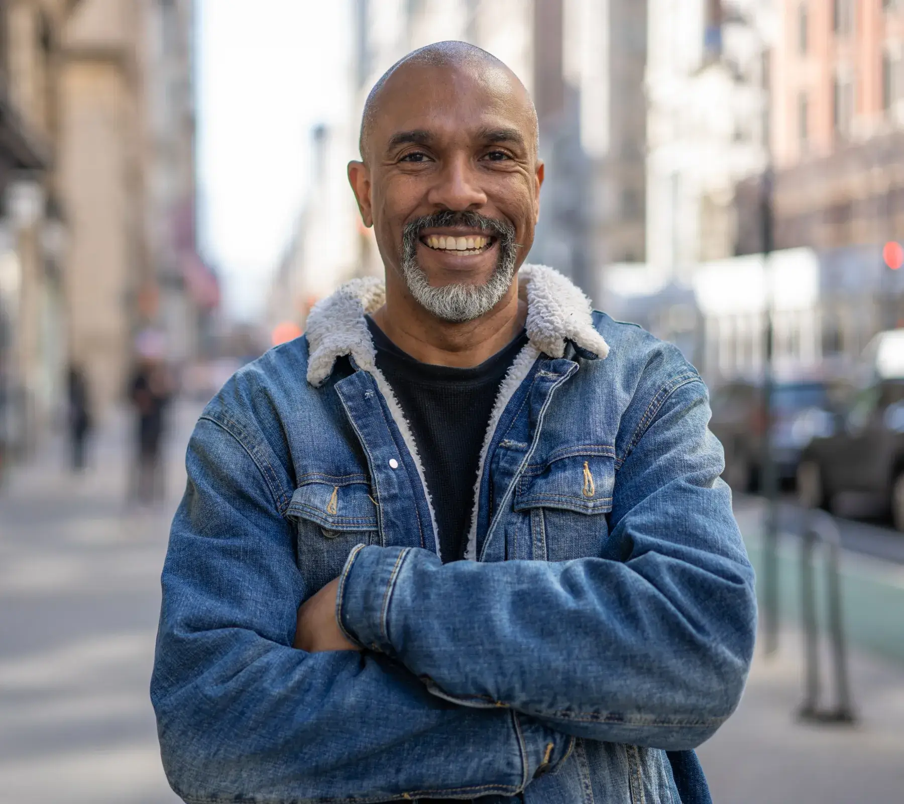 Smiling middle-aged man with a beard wearing a denim jacket with a shearling collar standing on a city street with arms crossed.