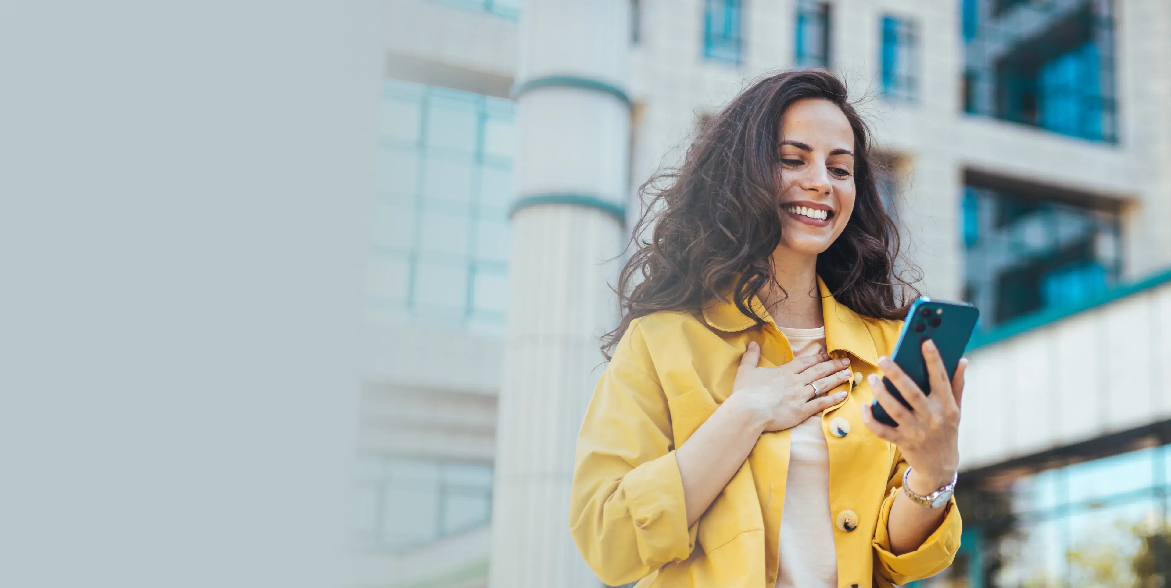 Smiling woman in yellow jacket looking at her smartphone outdoors near modern building.