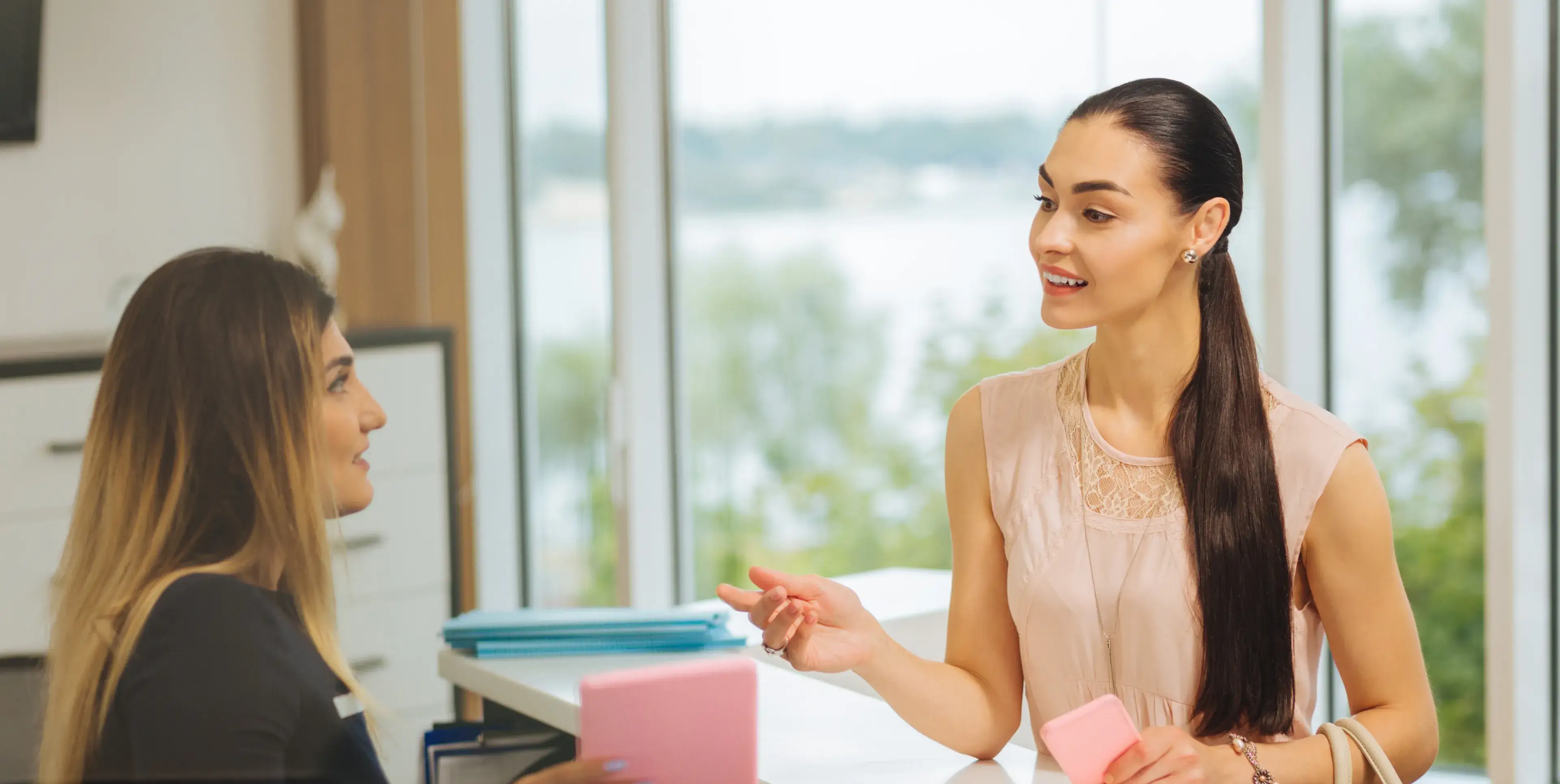 Two women engaged in a friendly conversation in a bright office setting, one holding a pink phone and the other seated with documents.