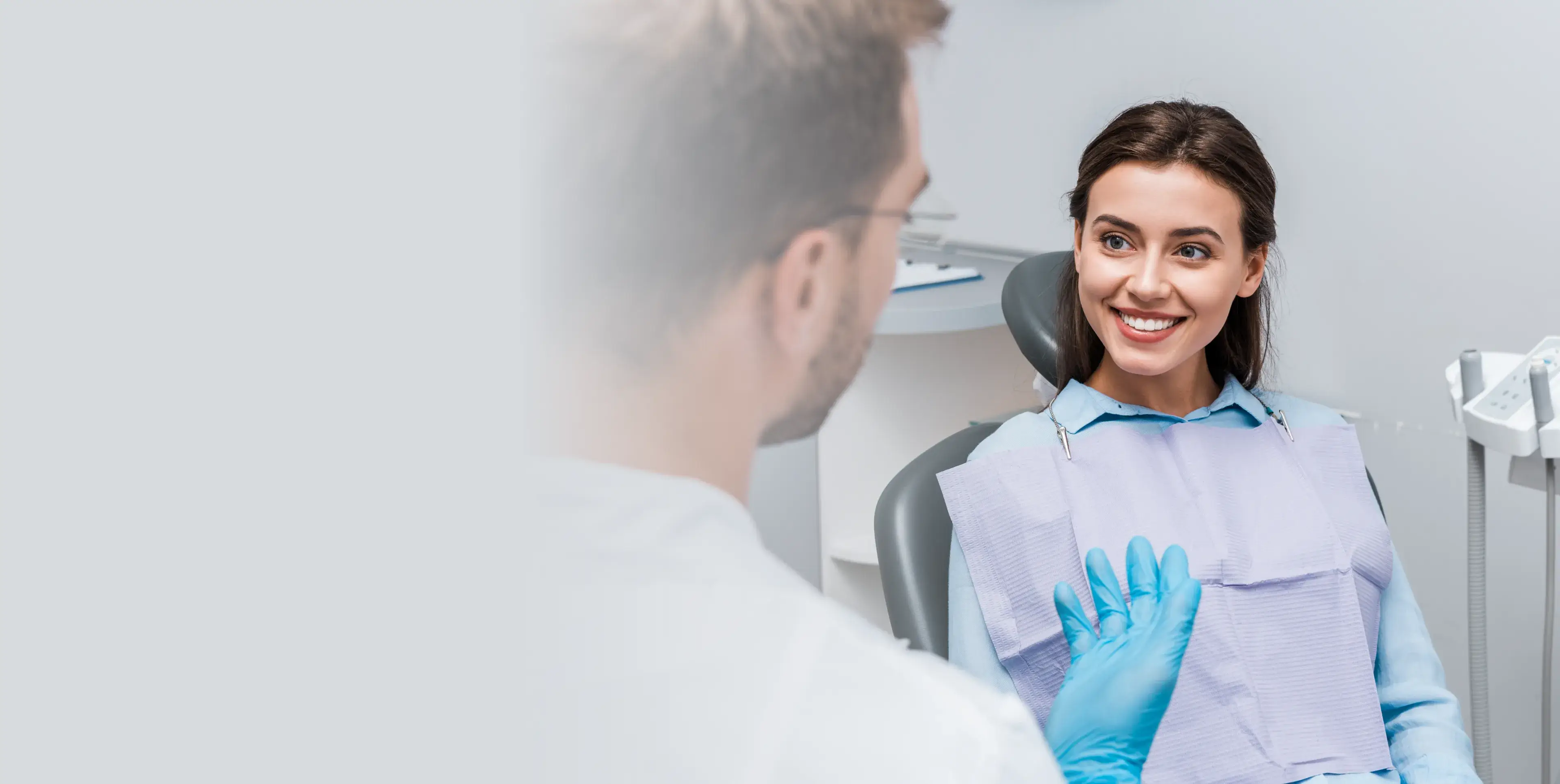 Smiling woman sitting in a dental chair talking to a dentist wearing blue gloves.