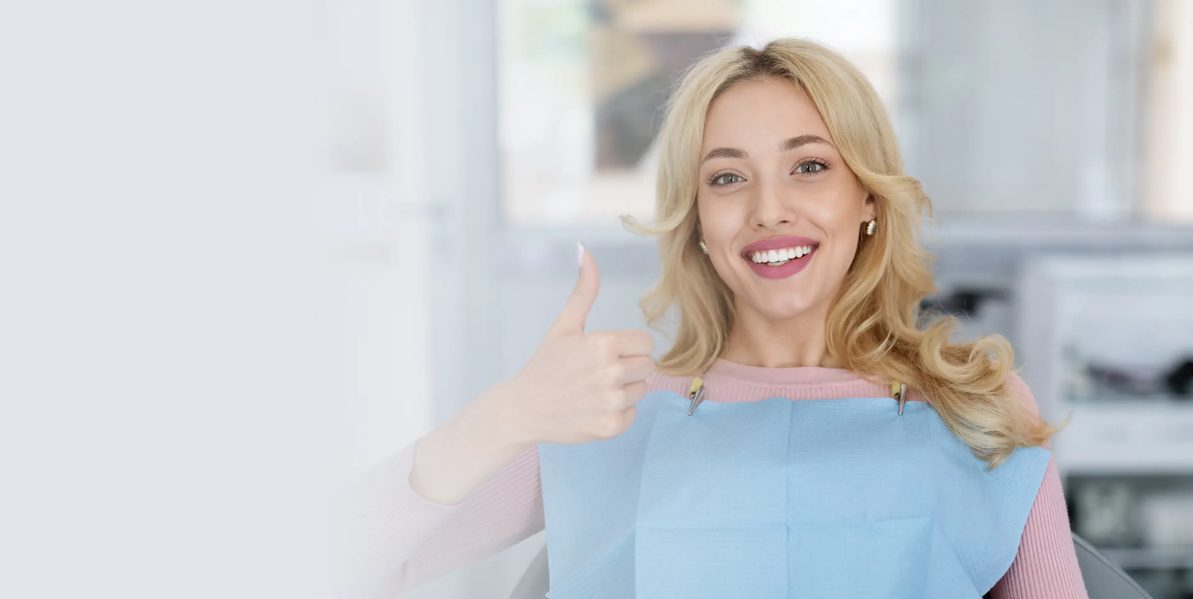 Smiling woman in a dental bib giving a thumbs up in a bright dental clinic.