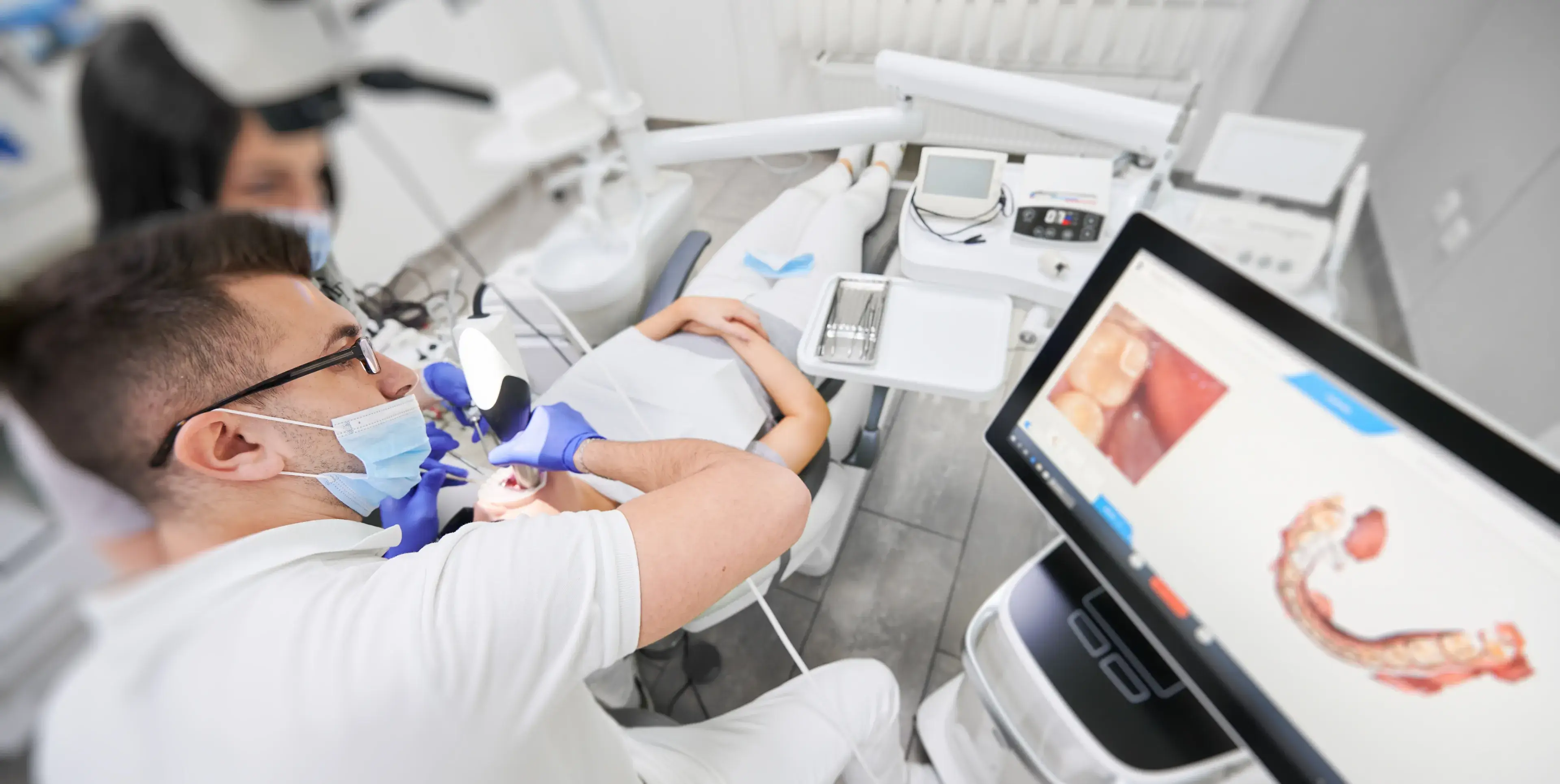 Dentist with mask and gloves examining a patient's teeth using a dental mirror and light, with a digital dental scan displayed on a nearby monitor.