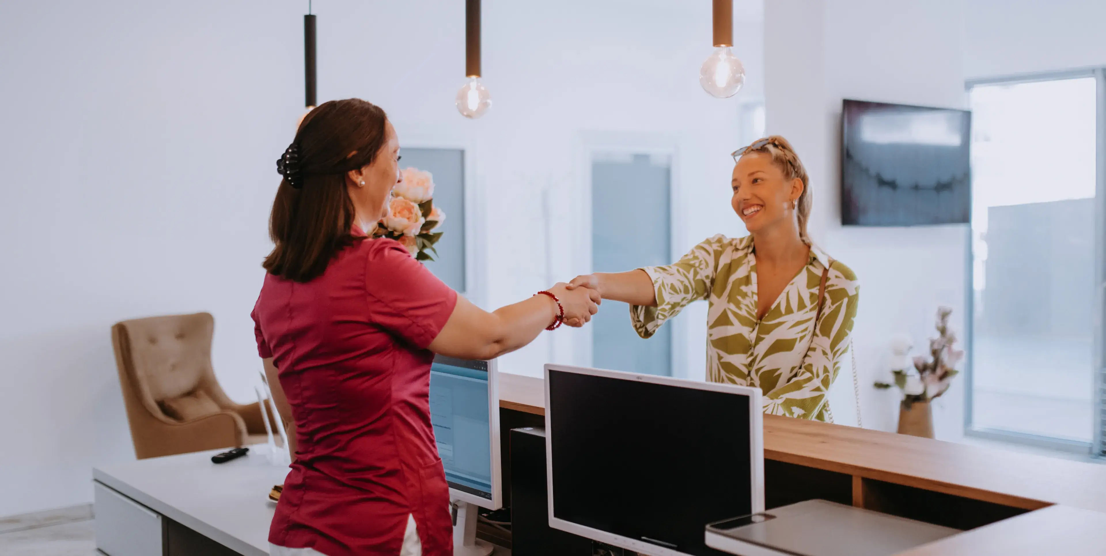 Smiling receptionist and visitor shaking hands across a reception desk in a bright office.