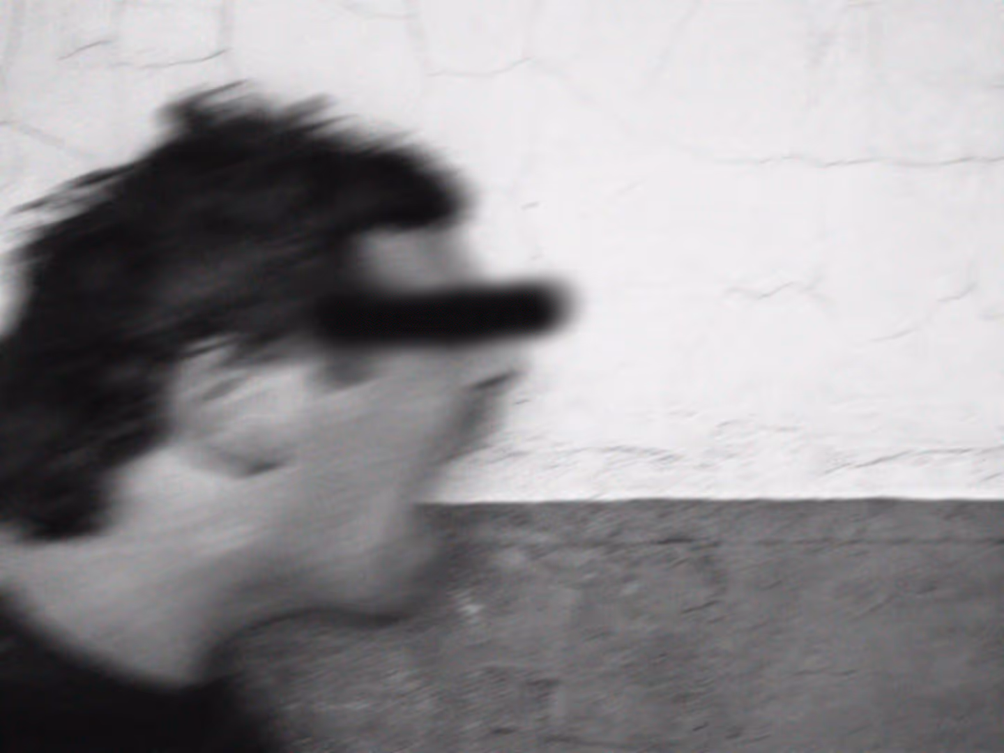 Black and white image of a man screaming in front of a white brick wall. A black bar has been spray painter to cover his eyes.