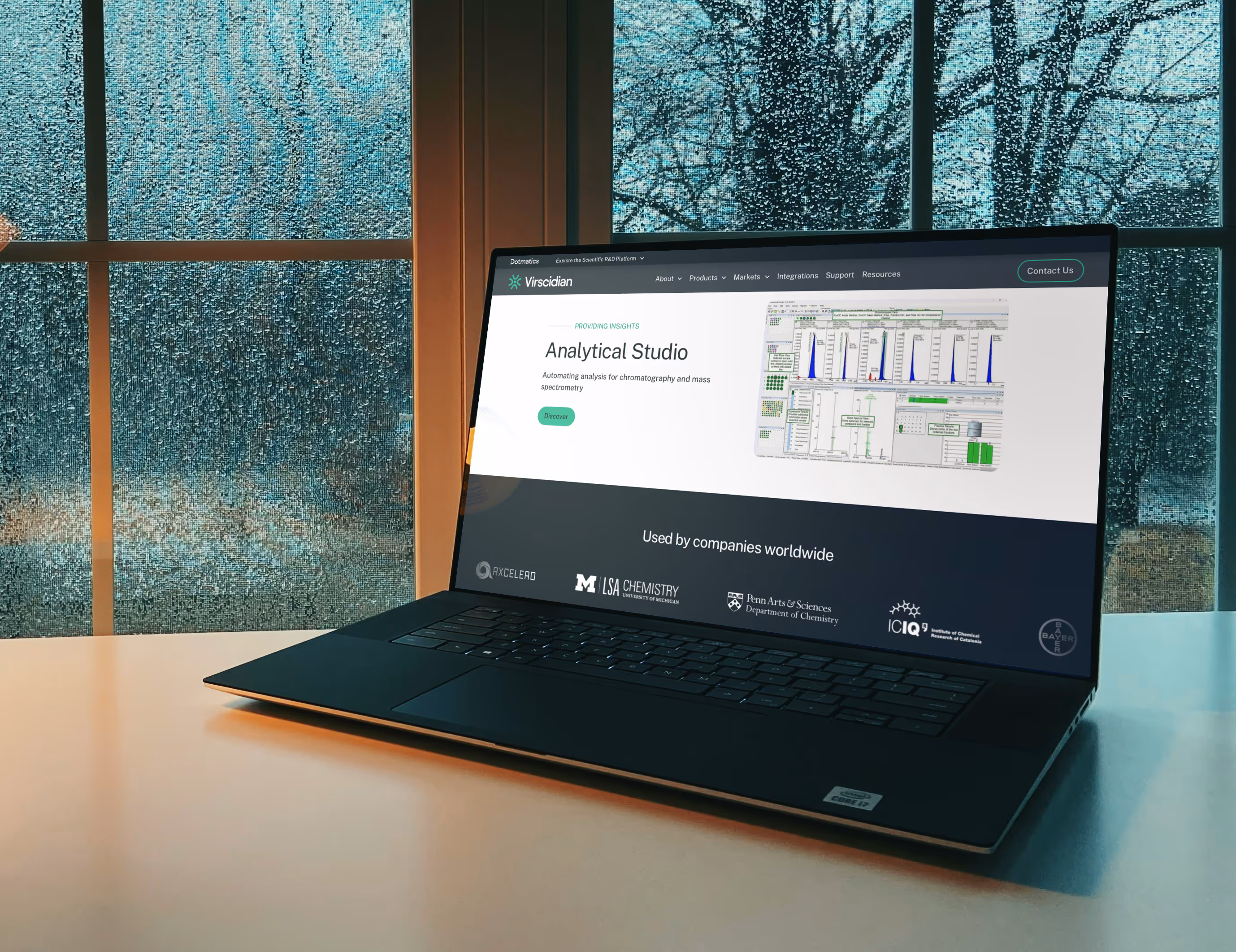 A laptop sitting on a desk in a warm home setting. Looking out the window behind the computer there is a cold snowy landscape. On the screen is the webpage for Virscidian.